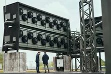 Zwei Personen stehen vor einer großen, industriellen Anlage mit mehreren Lüftern. Stahlträger und Rohre sind sichtbar.